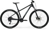 Side view of a black Merida BIG.NINE 100 mountain bike. It's a hardtail with front suspension, disc brakes, and black Maxxis tires with white lettering.