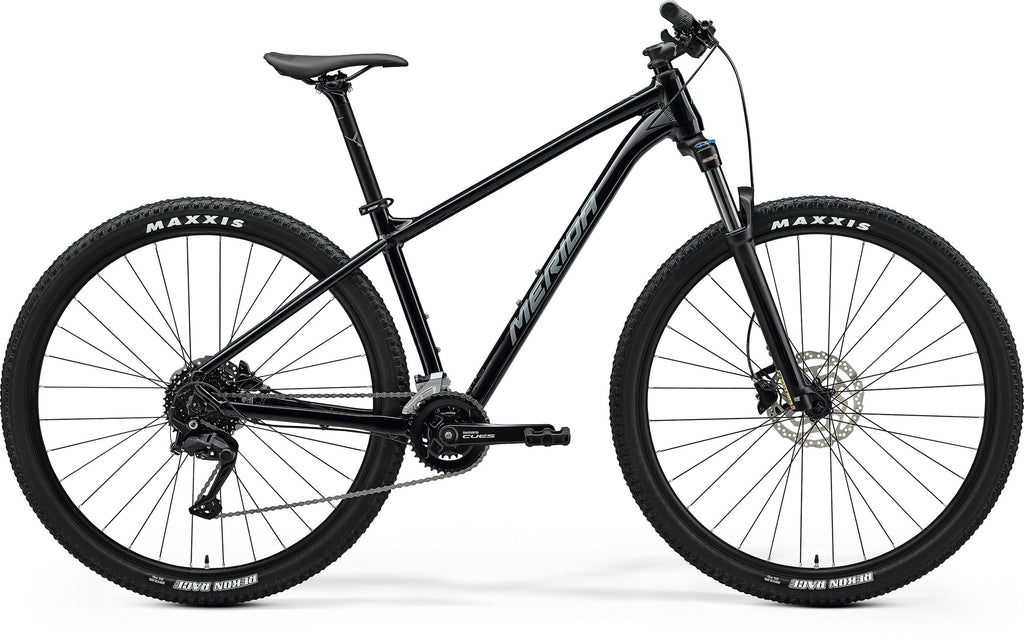Side view of a black Merida BIG.NINE 100 mountain bike. It's a hardtail with front suspension, disc brakes, and black Maxxis tires with white lettering.