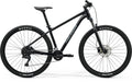 Side view of a black Merida BIG.NINE 100 mountain bike. It's a hardtail with front suspension, disc brakes, and black Maxxis tires with white lettering.