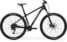 Side view of a black Merida BIG.NINE 100 mountain bike. It's a hardtail with front suspension, disc brakes, and black Maxxis tires with white lettering.