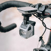 A close-up of the Magicshine EVO 1700 bike light mounted underneath bicycle handlebars, connected to a bike computer.