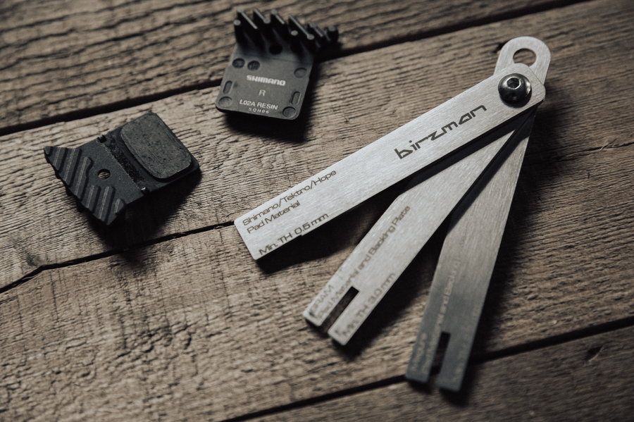 Birzman Brake Pad Wear Indicator laid beside worn Shimano resin brake pads, showing the tool in use for a wear check.