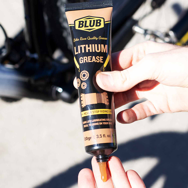 Blub Lithium Grease 100g tube being squeezed by hand next to a bicycle wheel and hub.
