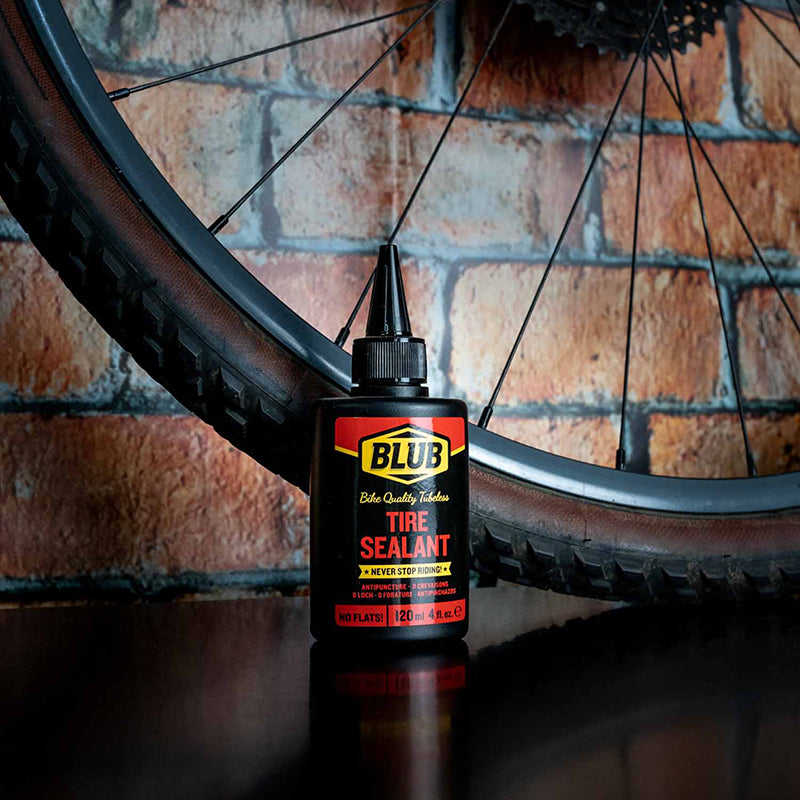 Blub Tire Sealant 120ml bottle standing beside a mountain bike wheel and tire against a brick wall.
