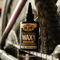 Blub Wax Lube 120ml bottle mounted on a bike frame near the chain and a knobby MTB tire.