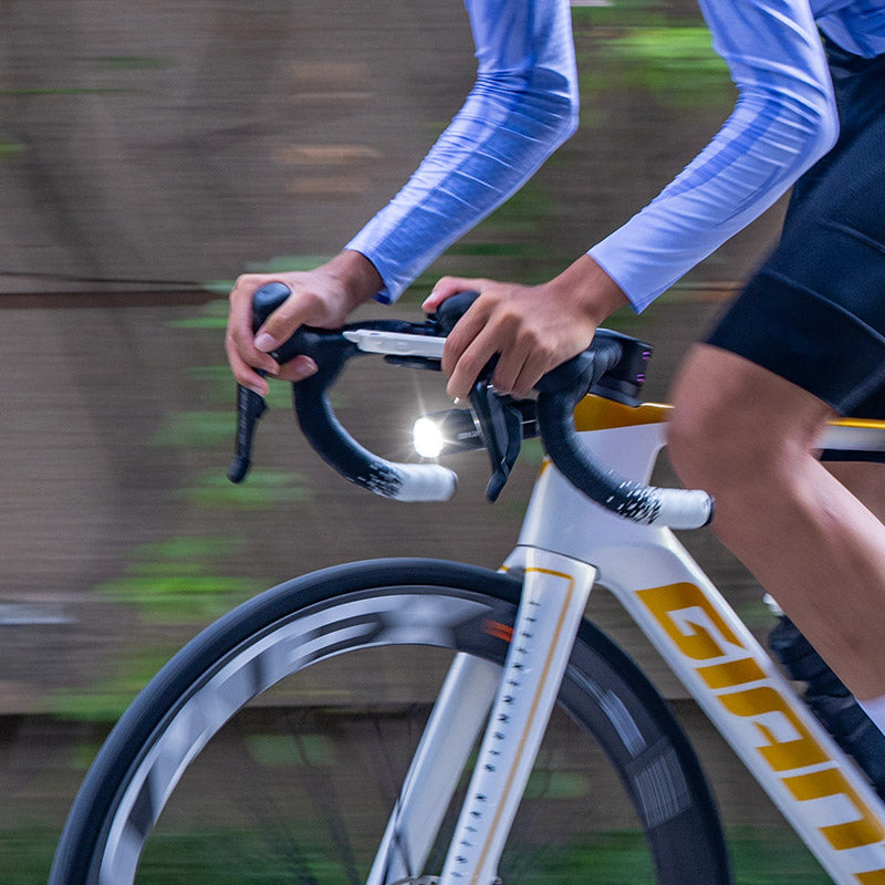 Close-up of a road cyclist's hands on drop handlebars with the Ravemen K1800 headlight lit during a ride.