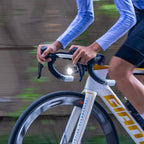 Close-up of a road cyclist's hands on drop handlebars with the Ravemen K1800 headlight lit during a ride.