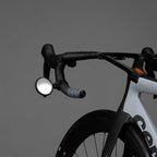A side view of a bicycle handlebar with the Guee i-See Safety Mirror mounted, showcasing its universal fit with a strong rubber strap.
